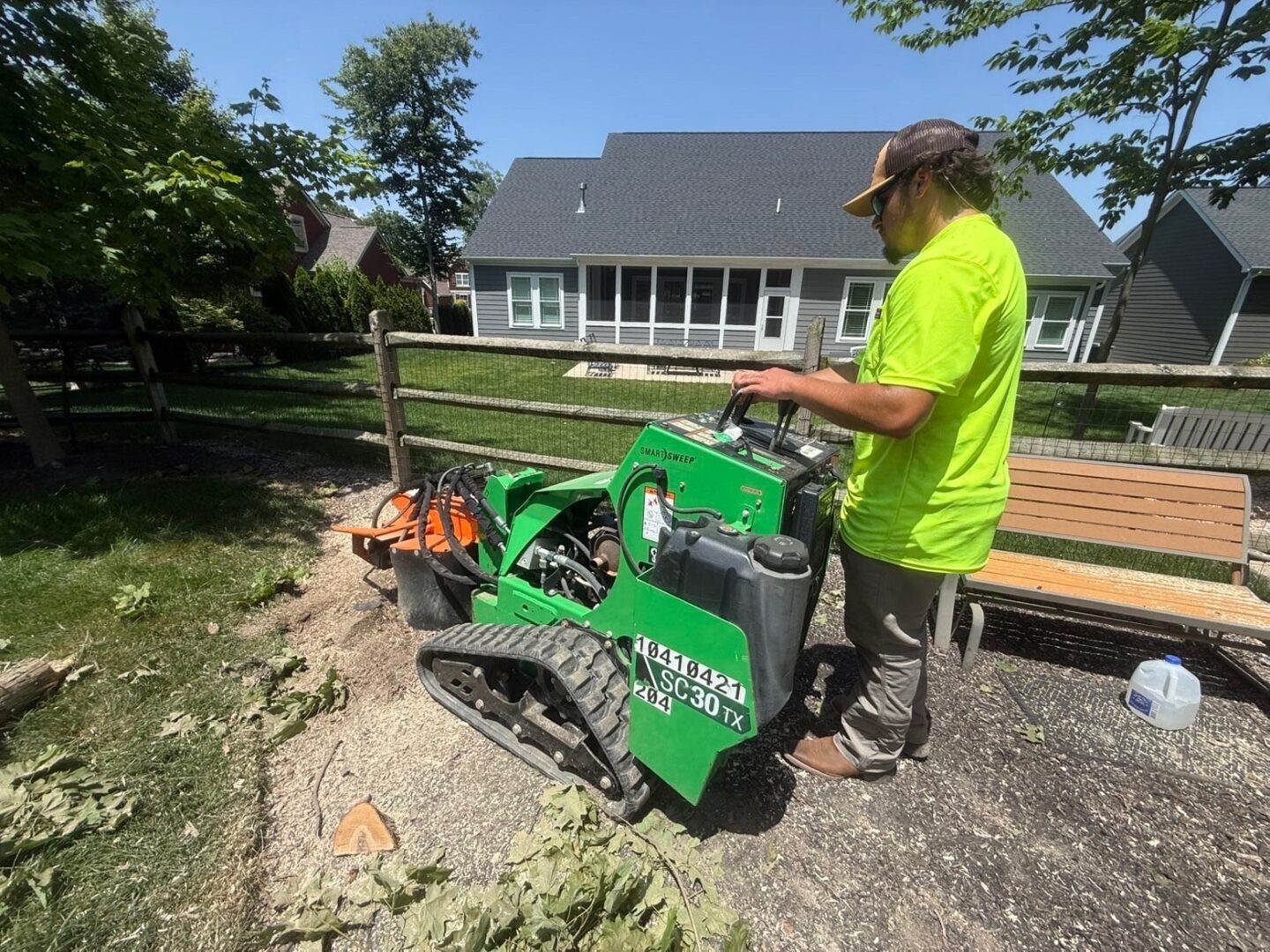 Crew member running a tracked walk-behind stump grinder on bare ground beside a wood fence in a back yard, with a house behind